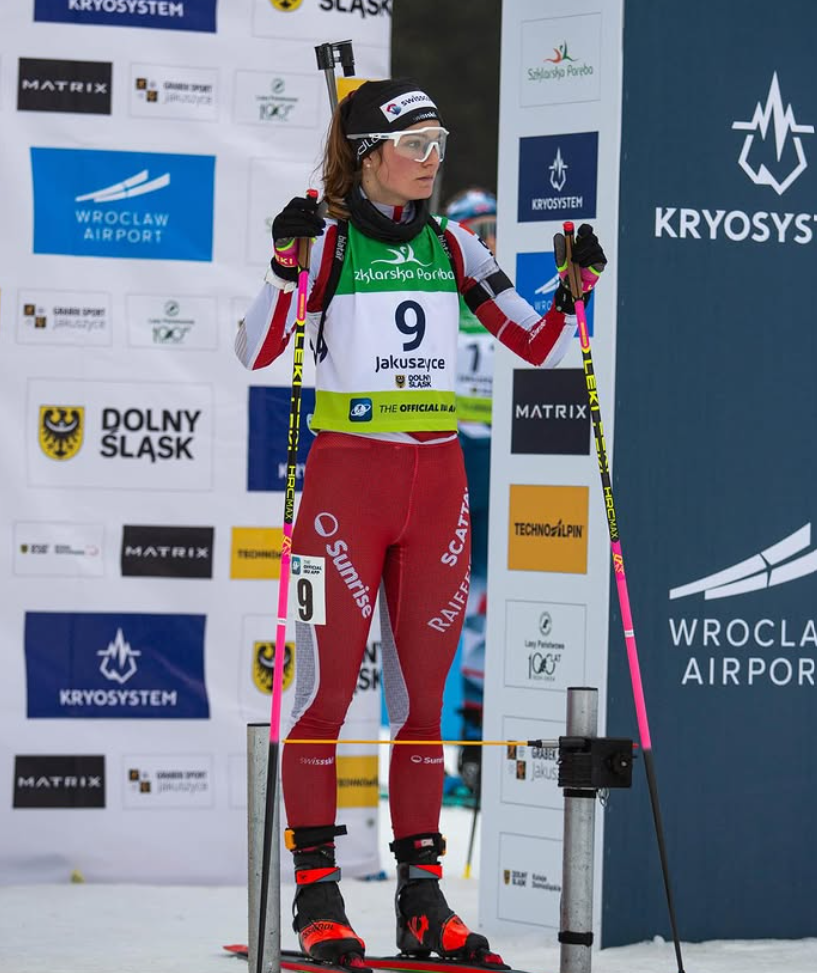 Eine Biathletin in rot-weißer Ausrüstung steht auf Skiern an der Startlinie und hält Skistöcke in der Hand. Sie trägt Helm und Schutzbrille. Im Hintergrund sind verschiedene Sponsorenlogos und Wintersportschilder zu sehen.