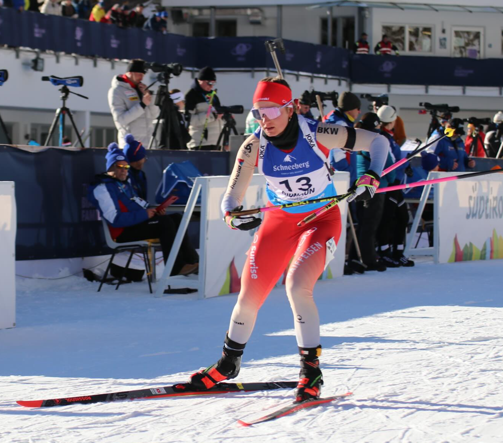 Ein Biathlet in rot-weißer Ausrüstung fährt auf einer verschneiten Strecke Ski, hält Skistöcke in der Hand und trägt die Startnummer 13, im Hintergrund sind Fotografen und Zuschauer.
