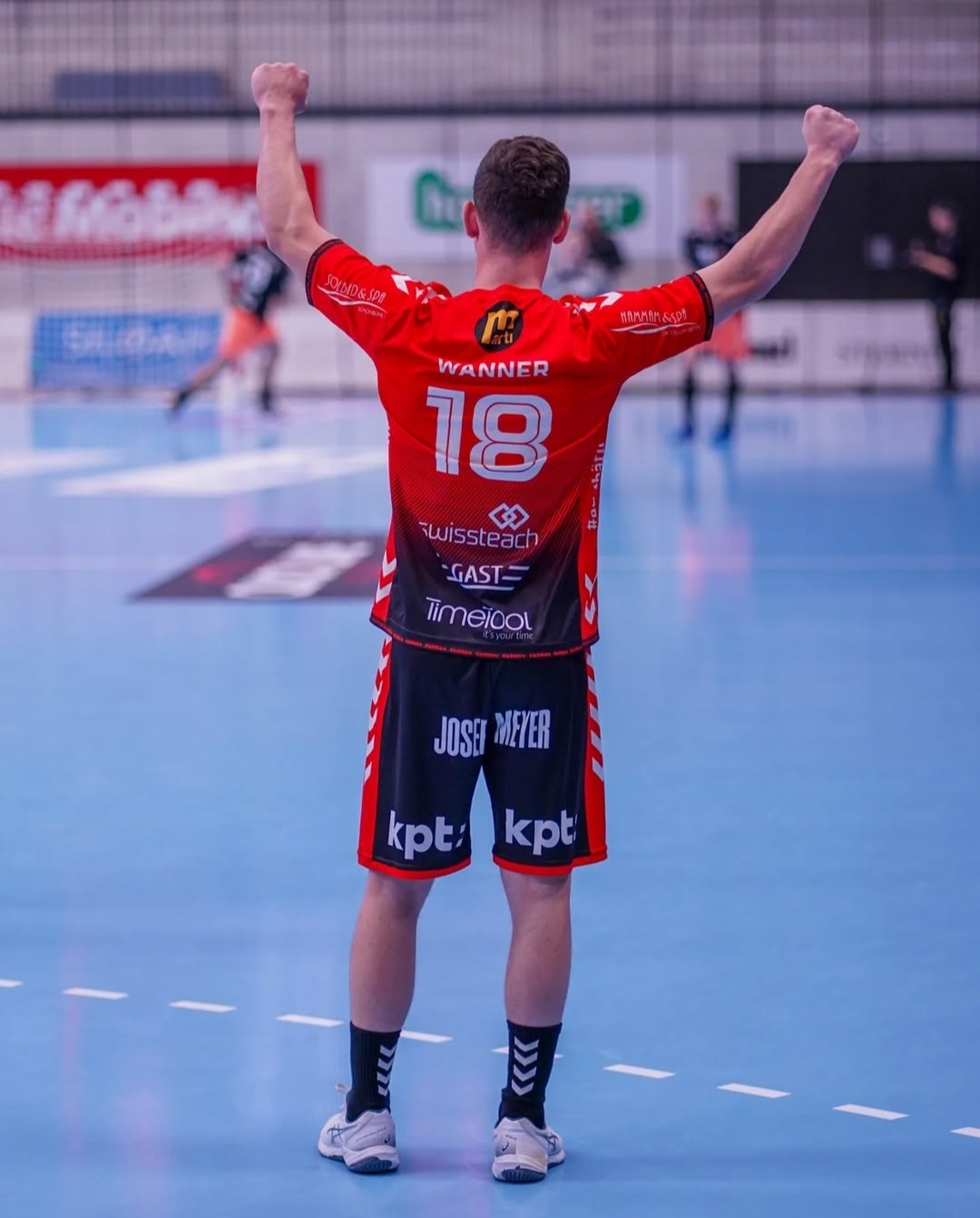 A male indoor athlete wearing a red jersey with the number 18 and “WANNER” on the back raises both arms in celebration on a blue court, with other players and blurred advertisements in the background.