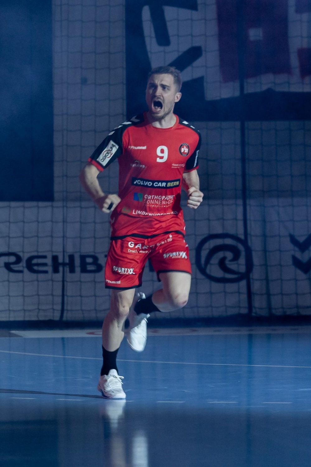 Simon Getzmann, ein männlicher Handballspieler in einer roten Uniform mit der Nummer 9, läuft auf einem Hallenplatz mit einem Netz im Hintergrund und wirkt aufgeregt und jubelnd.