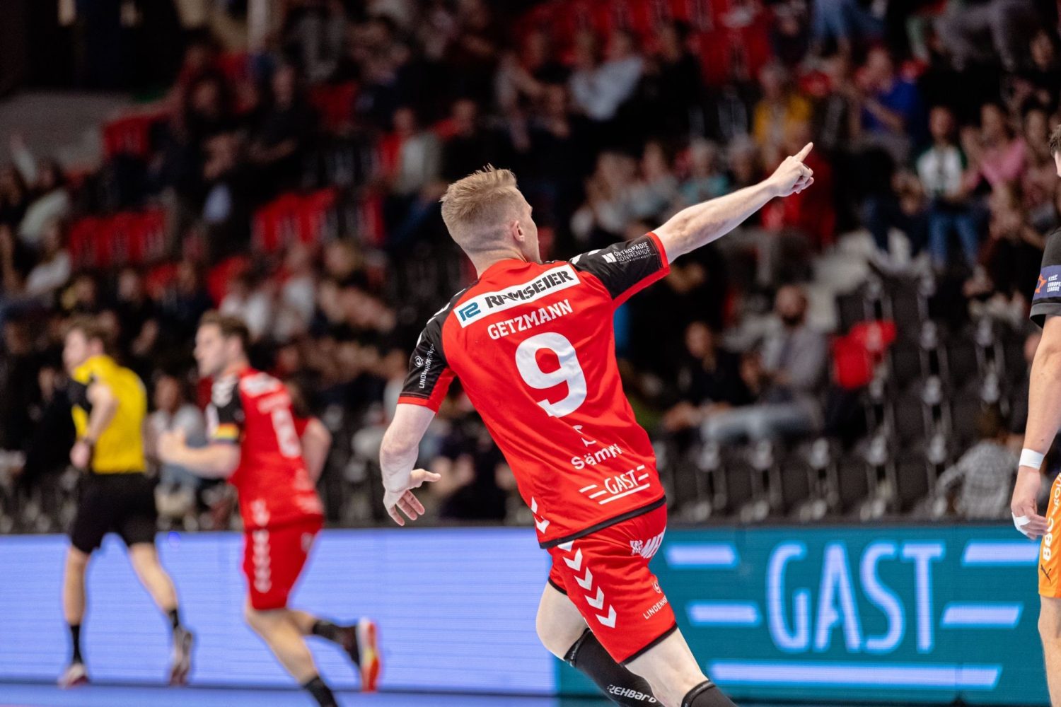 Handballspieler Simon Getzmann im roten Trikot mit der Nummer 9 streckt während eines Spiels seinen Arm nach vorne, während die Zuschauer im Hintergrund von der Tribüne aus zuschauen.