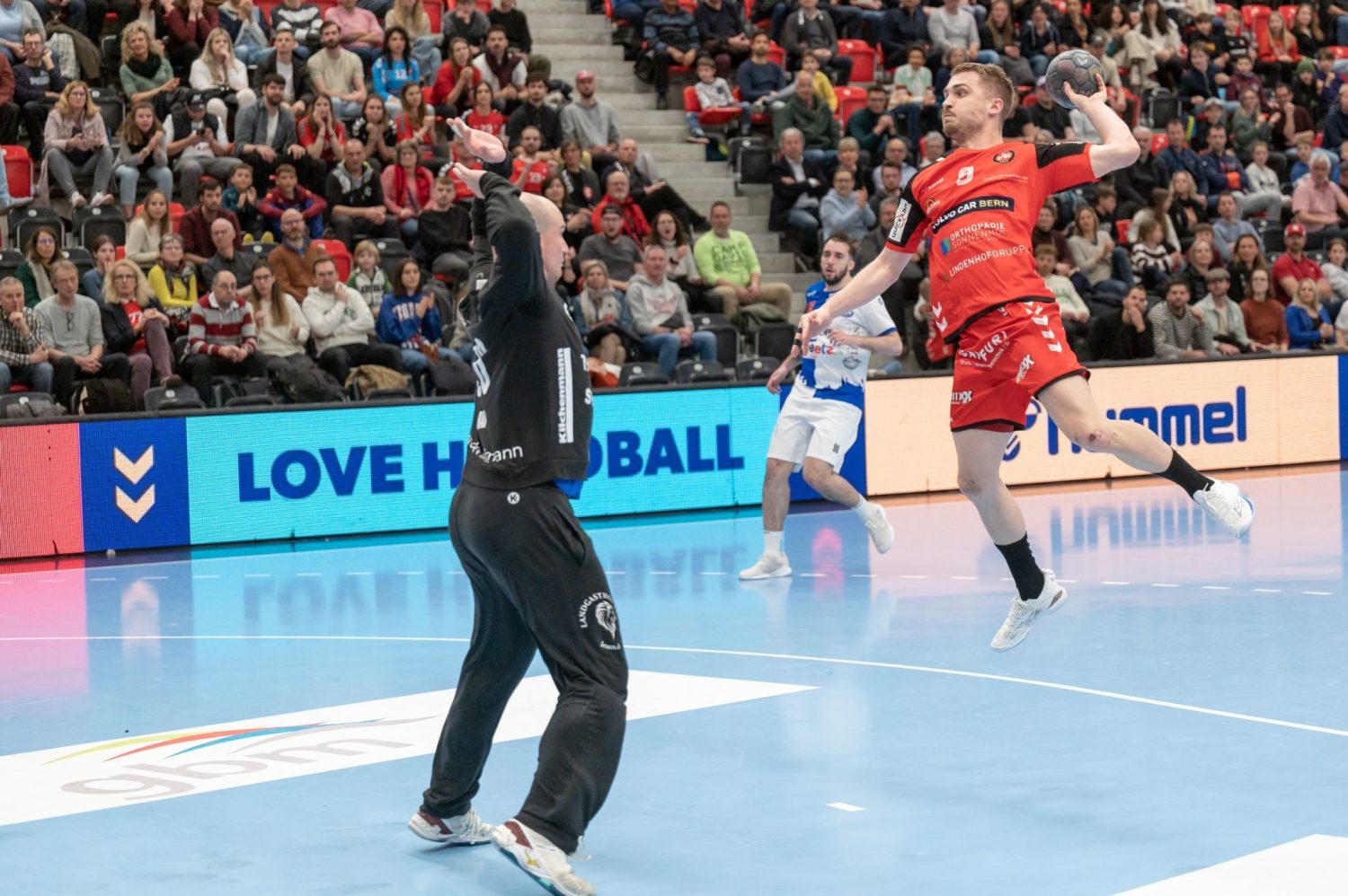 Handballspieler Simon Getzmann in roter Uniform springt hoch, um den Ball zu schießen, während ein Torwart in Schwarz die Arme zum Blocken hebt. Die Zuschauer auf der Tribüne schauen zu.