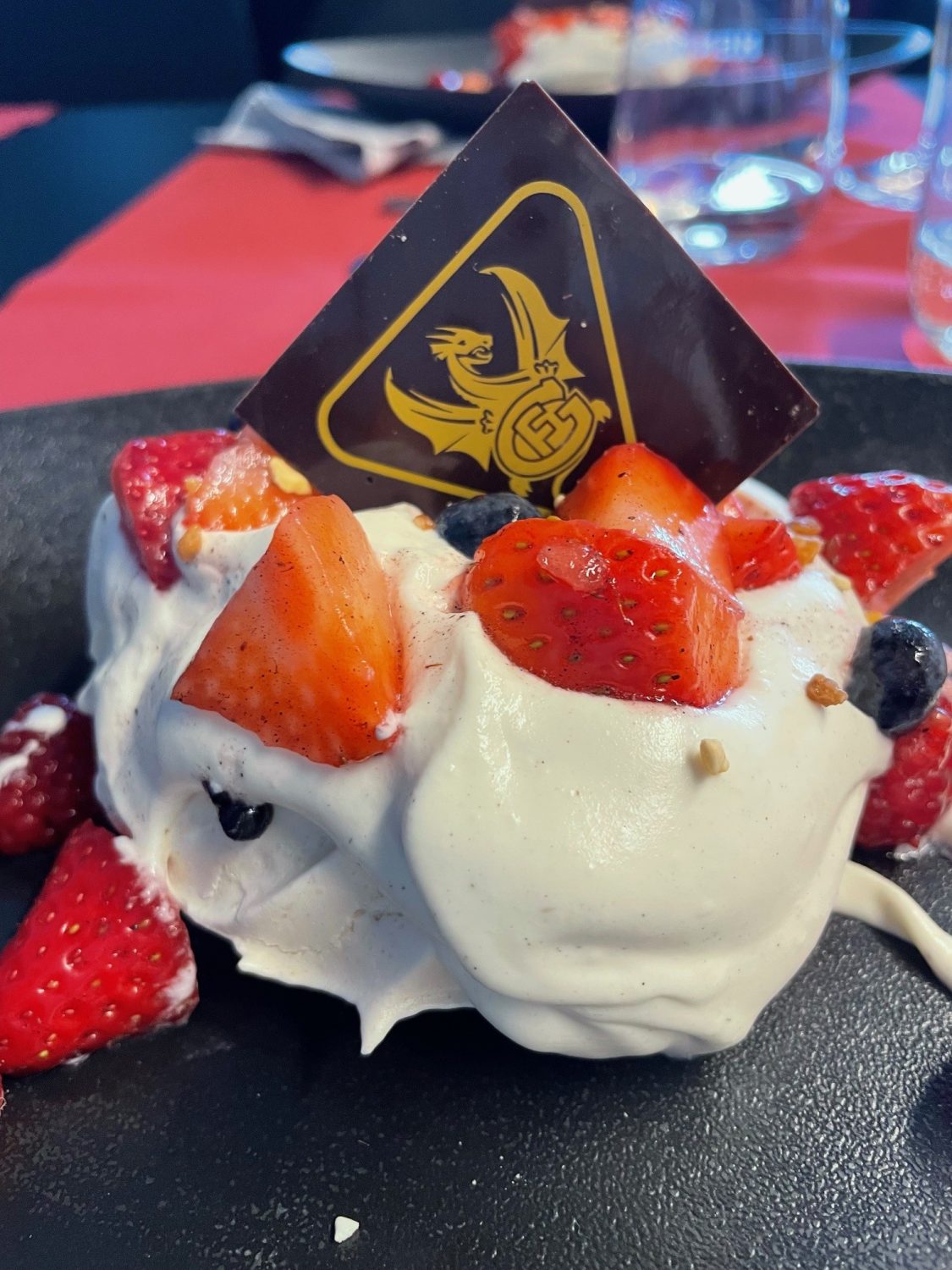 Ein Dessert mit Schlagsahne, Erdbeeren, Blaubeeren und einem quadratischen Stück dunkler Schokolade mit dem Logo des HC Fribourg-Gottéron, serviert auf einem dunklen Teller mit Gläsern und roten Tischakzenten im Hintergrund.