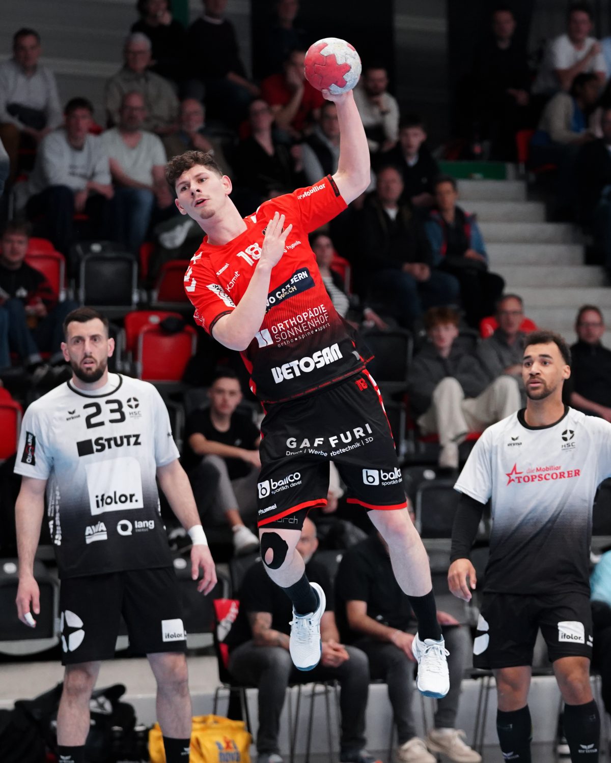 A handball player in a red jersey jumps mid-air to throw the ball, while two opposing players in white jerseys watch. The action takes place in an indoor arena with spectators in the background.