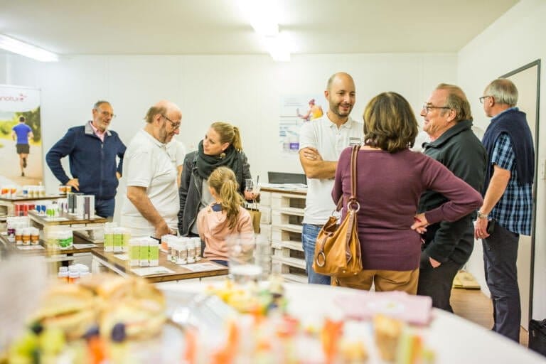 Eine Gruppe von Menschen, darunter Erwachsene und ein Kind, versammelt sich und unterhält sich an Tischen, auf denen Gläser und Produkte in einem hell erleuchteten Innenraum ausgestellt sind, was auf einen Tag hinter der offenen Tür hindeutet, wo sich Besucher anmelden können.