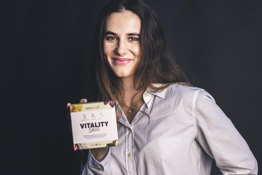 Talina Gantenbein, mit langen braunen Haaren und einem weißen Hemd mit Knöpfen, lächelt, während sie vor einem dunklen Hintergrund eine Schachtel mit der Aufschrift „Vitality Shot“ hält.
