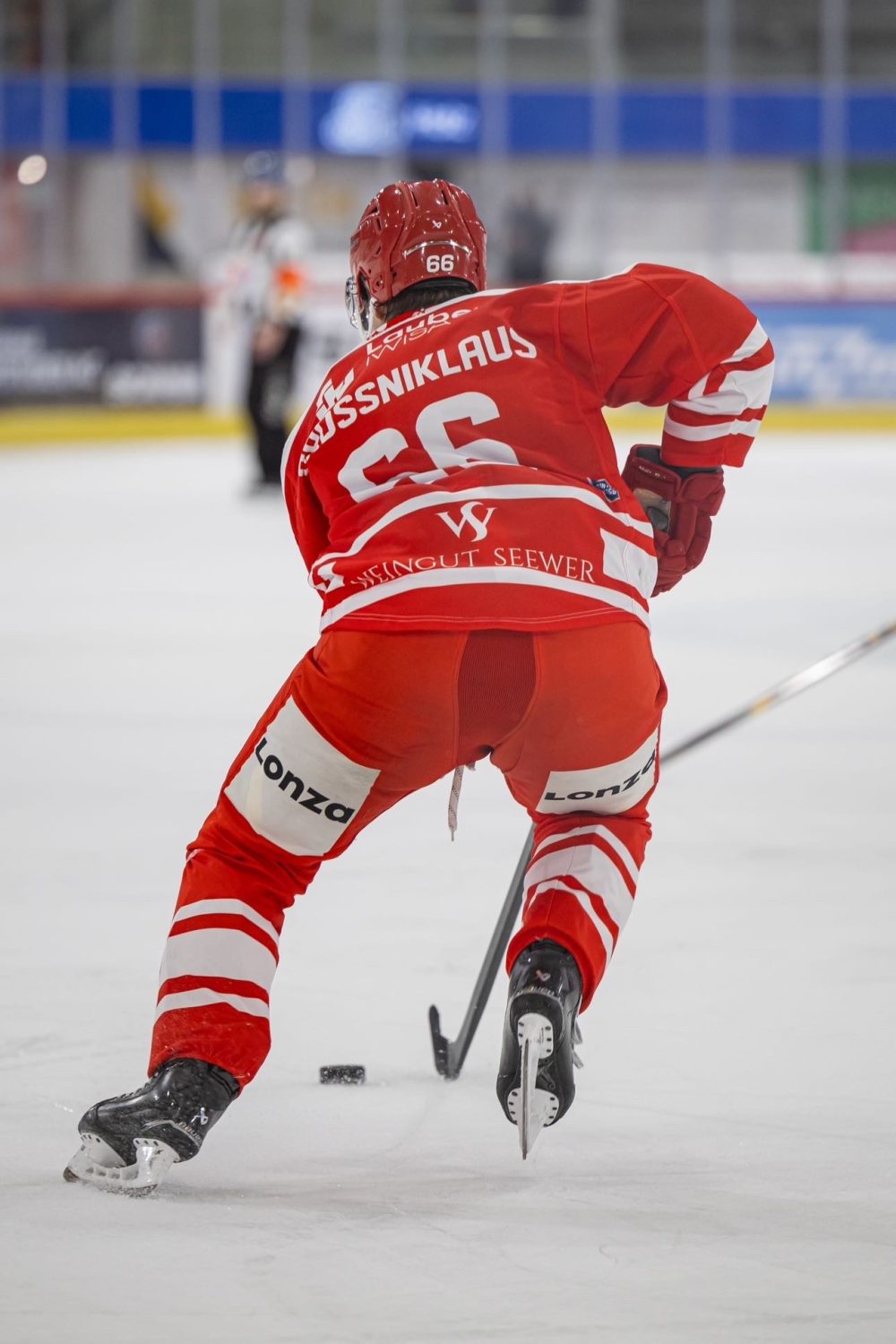 A hockey player in a red and white uniform, number 66, skates on the ice with a hockey stick, preparing to play the puck. The player’s name, “SÜSSNIKLAUS,” is visible on the back of the jersey.