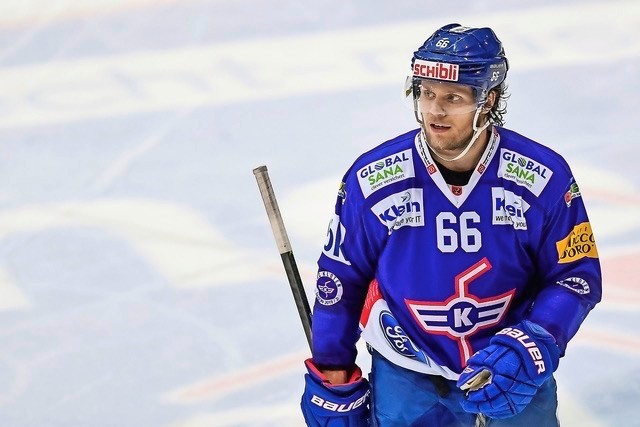 A male ice hockey player in a blue uniform and helmet, number 66, stands on the ice rink holding a hockey stick. His jersey features sponsor logos and a red, white, and blue team emblem.