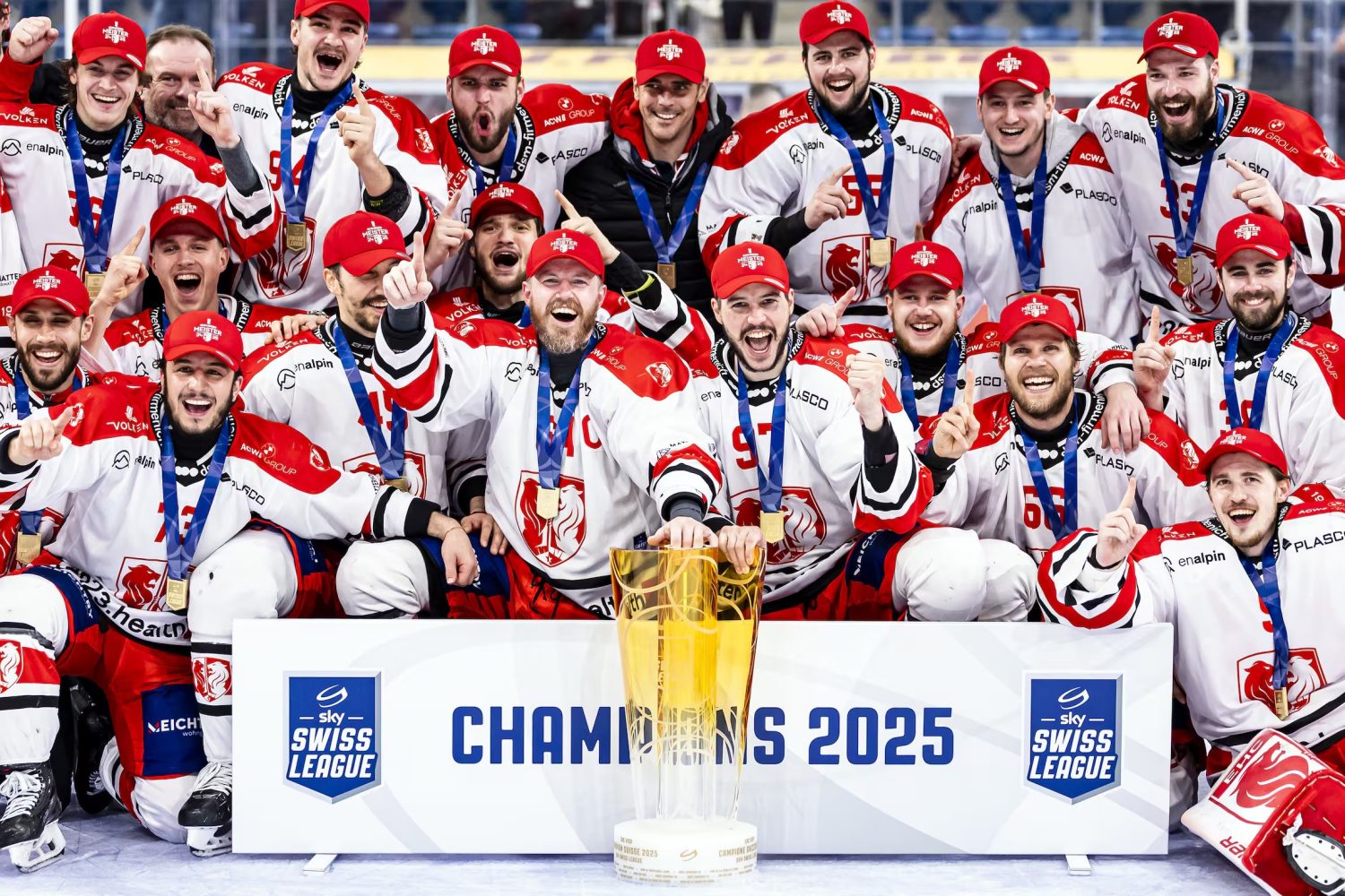 A jubilant ice hockey team in white and red uniforms celebrates with gold medals around their necks, gathered behind a large gold trophy and a sign that reads CHAMPIONS 2025 at the Swiss League.