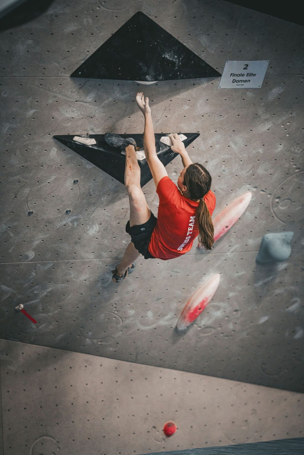 Valentina von Gunten, in rotem Shirt und schwarzen Shorts, klettert eine Indoor-Kletterwand hoch und umklammert mit Händen und Füßen schwarze Dreiecksgriffe. Ihre langen Haare sind zu einem Pferdeschwanz zurückgebunden.