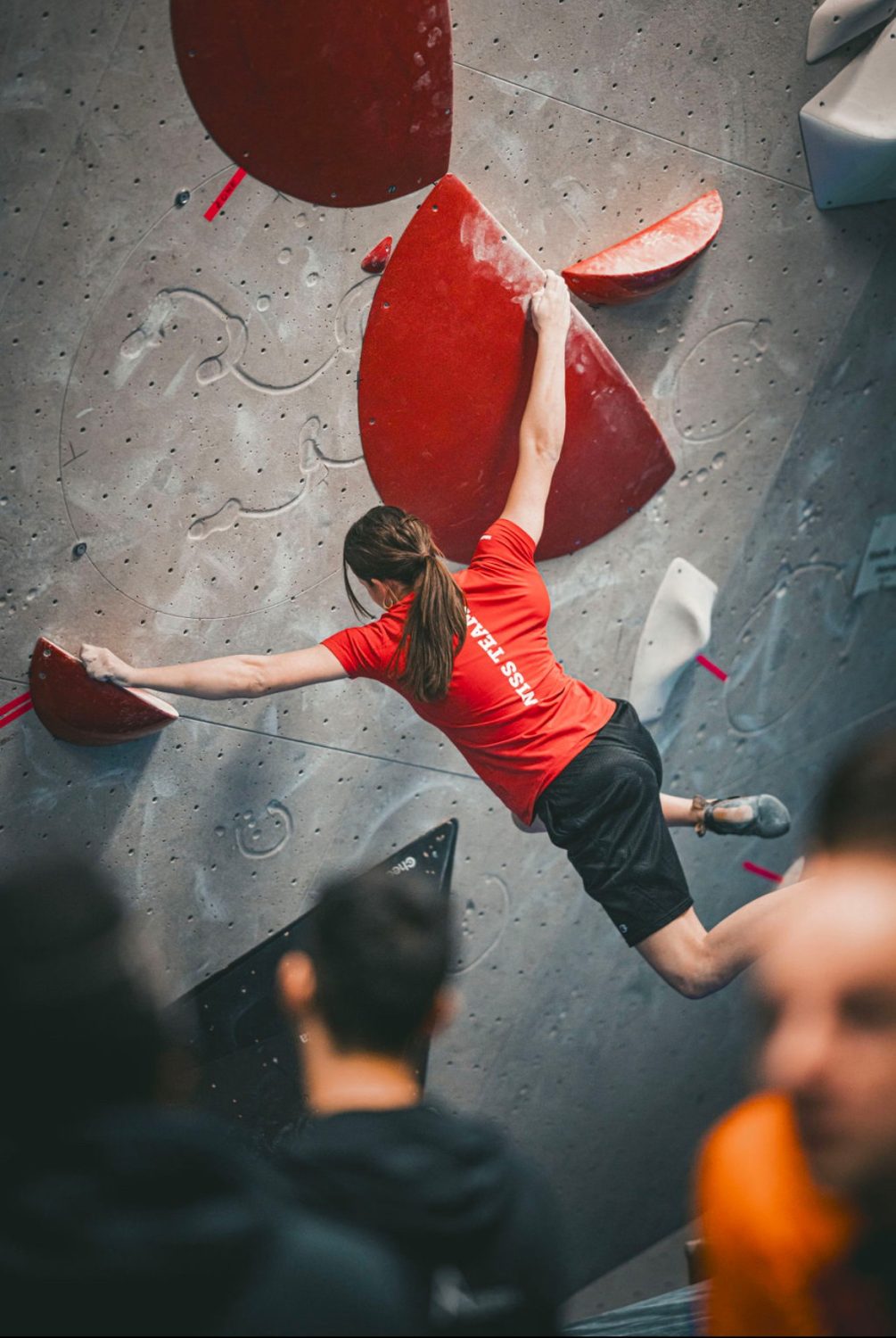 Valentina von Gunten, in rotem Shirt und schwarzen Shorts, klettert eine Indoor-Boulderwand hoch und greift dabei nach großen roten Griffen. Mehrere Personen schauen von unten zu, leicht unscharf.
