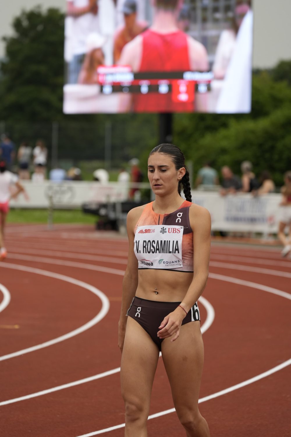A female athlete in a brown and orange uniform walking on a running track, looking focused. Her bib reads V. Rosamilia. A large screen with blurred figures is visible in the background.