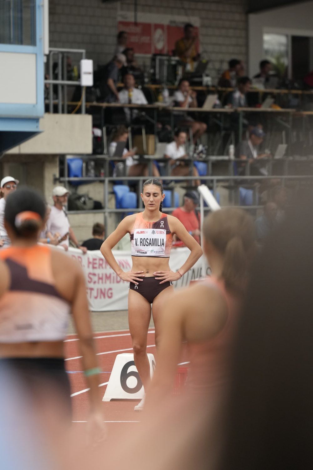 A female track athlete wearing a race bib that reads V. ROSAMILIA stands on a running track near the starting line, appearing focused, with other athletes and spectators visible in the background.