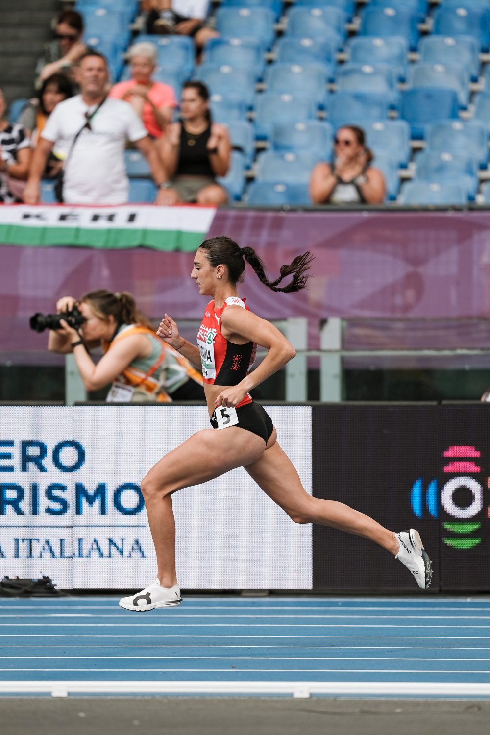 A female athlete in a running uniform sprints on a blue track at a stadium, with spectators and a photographer visible in the background.