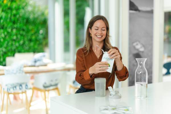 ageaway2 Eine lächelnde Frau sitzt an einer hellen Küchentheke und öffnet eine Packung „AG1“ von kingnature Nahrungsergänzung aus der Schweiz neben einer Glaskaraffe mit Wasser, einer Shaker-Flasche und anderen Packungen mit Nahrungsergänzungsmitteln.