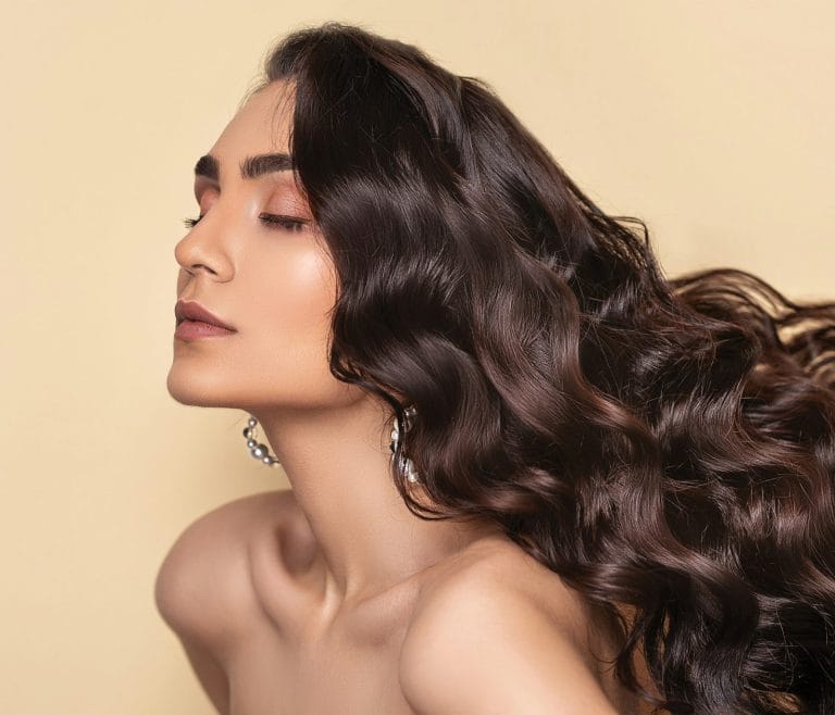 Eine Frau mit langem, welligem, dunklem Haar posiert vor einem hellbeigen Hintergrund, die Augen geschlossen, und präsentiert glatte Haut und elegante Ohrringe – ihr üppiges Haar spiegelt die Vorteile von Biotin wider.