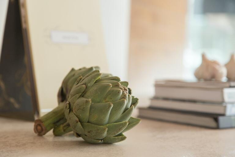 Eine frische Artischocke liegt auf einer hellen Oberfläche mit Büchern im Hintergrund und schafft eine gemütliche Atmosphäre, die auf subtile Weise die Essenz von Amaro Vida heraufbeschwört.