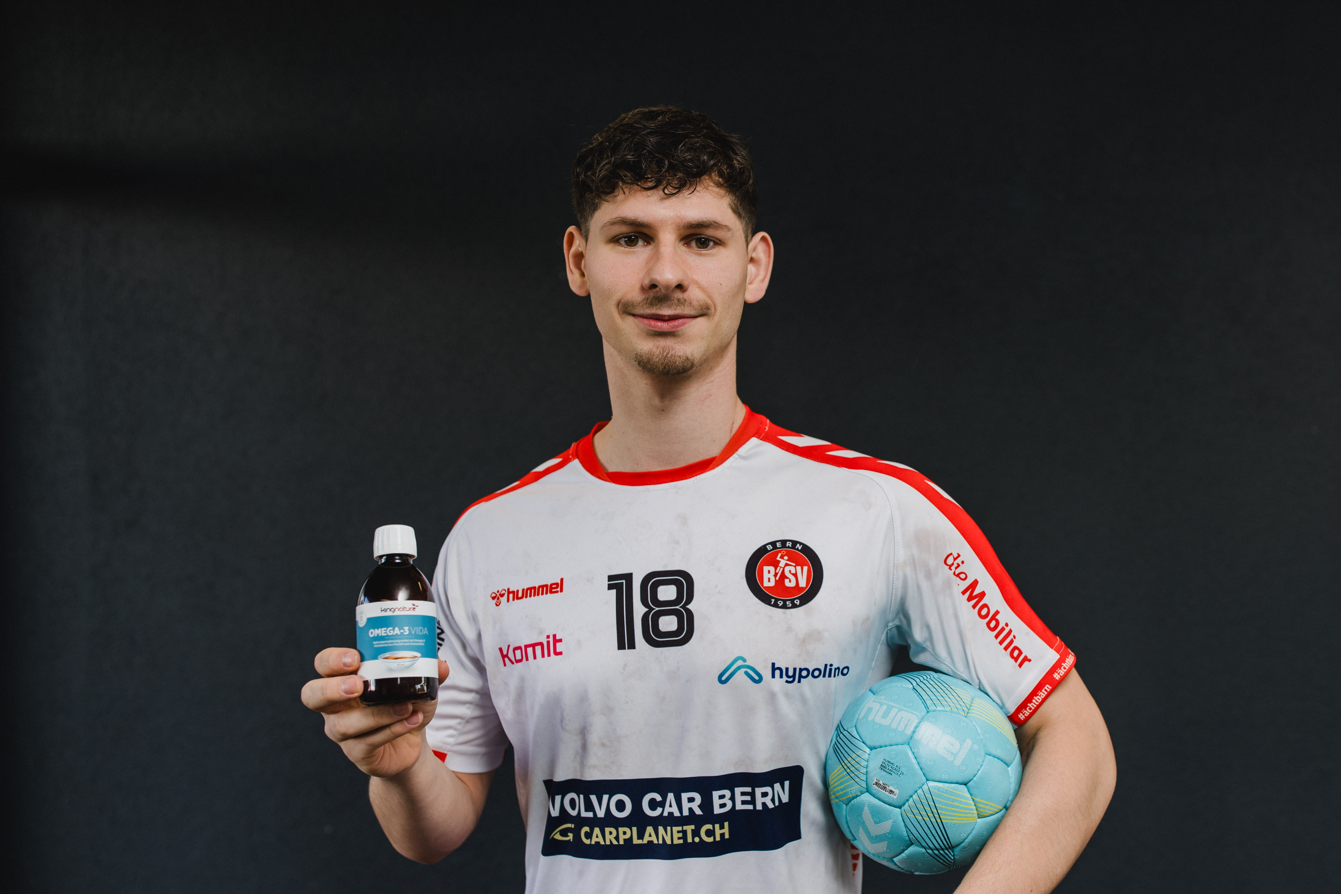 Levin Wanner, ein männlicher Sportler in einem weiß-roten Sporttrikot mit der Nummer 18, hält vor einem dunklen Hintergrund eine blaue Sportflasche in der einen und einen blauen Handball in der anderen Hand.