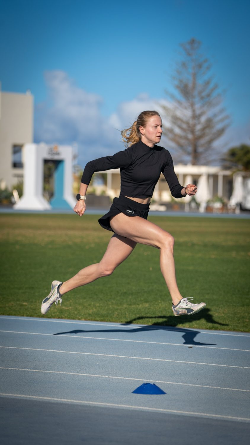 Linda Bichsel läuft in Sportkleidung in der Luft auf einer Laufbahn, mit einem Bein nach vorne und einem nach hinten, an einem sonnigen Tag mit blauem Himmel und grünem Gras im Hintergrund.