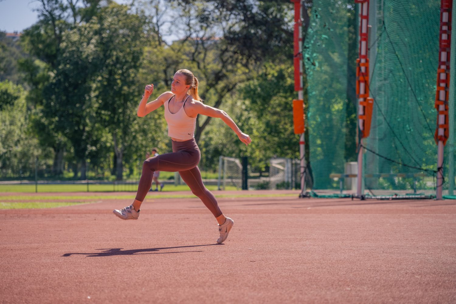 Linda Bichsel läuft in Sportkleidung auf einer roten Laufbahn im Freien, umgeben von grünen Bäumen und einem Sportnetz, wobei das Sonnenlicht Schatten auf den Boden wirft.
