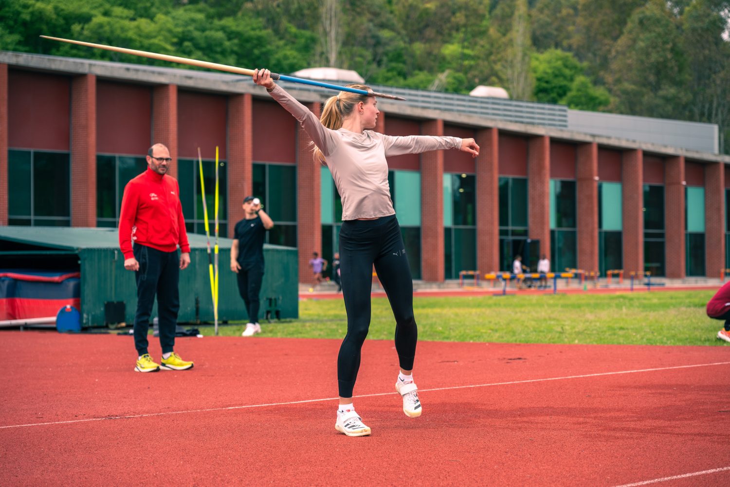 Linda Bichsel bereitet sich in Sportkleidung auf einem Leichtathletikfeld auf einen Speerwurf vor, während Trainer und Athleten im Hintergrund in der Nähe eines Backsteingebäudes zuschauen.