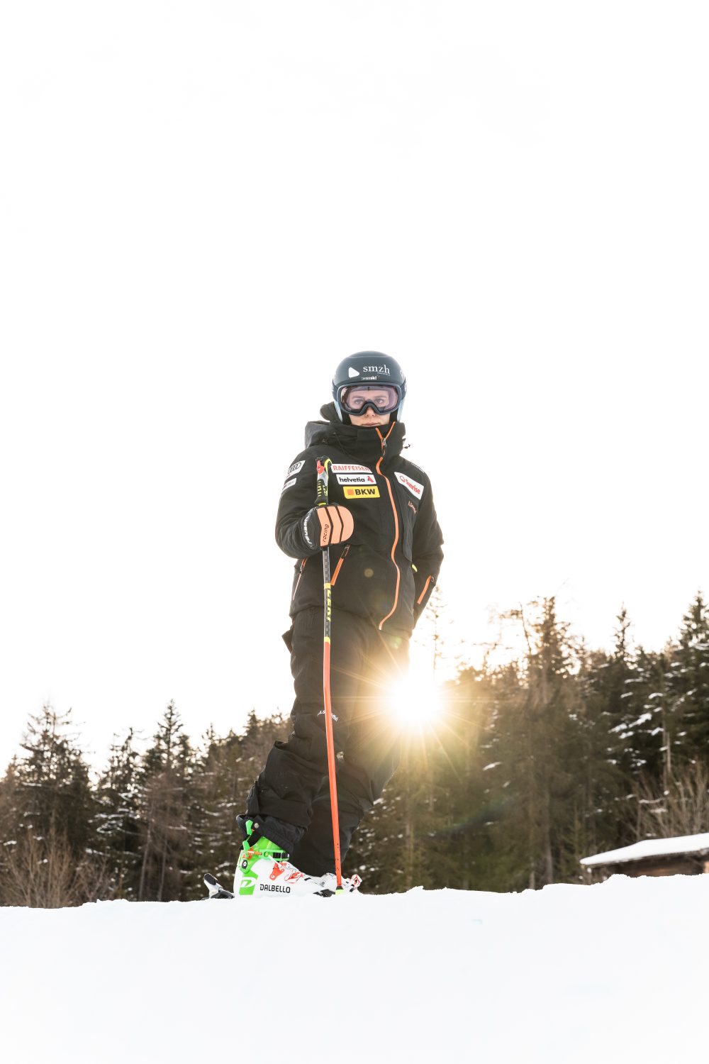 A skier dressed in black winter gear and goggles stands on snow with trees in the background and the sun shining behind their legs. The skier holds ski poles and wears green and white ski boots.