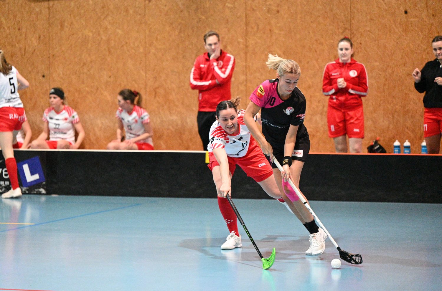 Two women compete for the ball in a floorball game; one in a black and pink uniform controls the ball while the other in a red and white uniform attempts to intercept. Teammates and coaches watch from the background.
