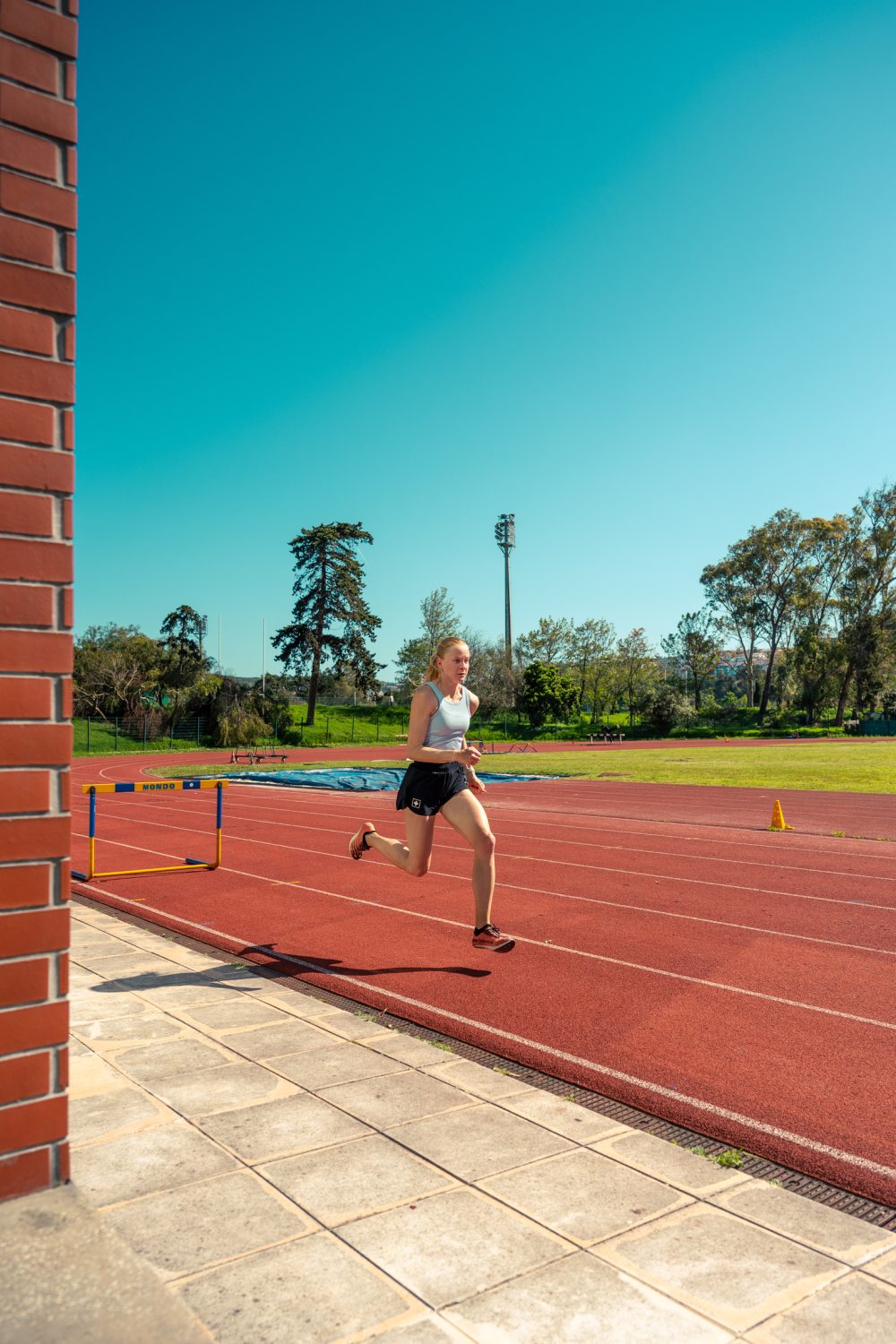 Linda Bichsel läuft in Sportkleidung auf einer roten Bahn und springt in der Luft über eine Hürde, im Hintergrund sind Bäume und ein klarer blauer Himmel zu sehen.