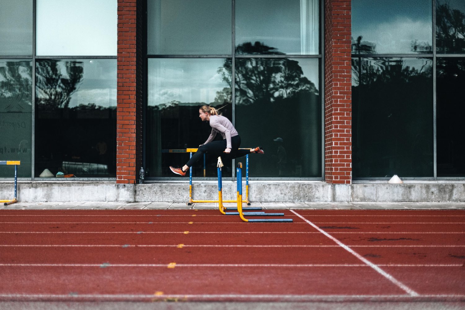 Linda Bichsel springt in Sportkleidung über eine Hürde auf einer Außenbahn, im Hintergrund große Fenster und ein Backsteingebäude.