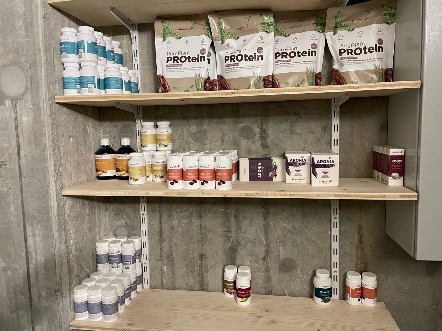Regale mit Flaschen, Gläsern und Beuteln des PurePlant-Proteinpulvers sind ordentlich in Reihen auf Holzregalen angeordnet. Die Ausstellung befindet sich in einem Raum mit Betonwänden und erzeugt so ein klares Erscheinungsbild, das an eine Schweizer Eishockey-Umkleidekabine erinnert.