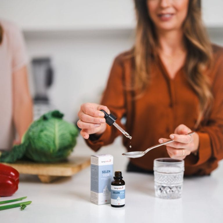 Eine Frau gibt mit einer Pipette Selen Vida Tropfen aus einer braunen Flasche auf einen Löffel, im Hintergrund auf der Küchentheke stehen ein Glas Wasser, Gemüse und eine weitere Person.