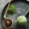 Auf einem schwarzen Teller liegen eine Kugel grünes Matcha-Eis, ein Bambusbesen, ein Löffel und Matcha-Pulver – ideal für alle, die Matcha Pulver Bio kunstvoll arrangiert kaufen möchten.
