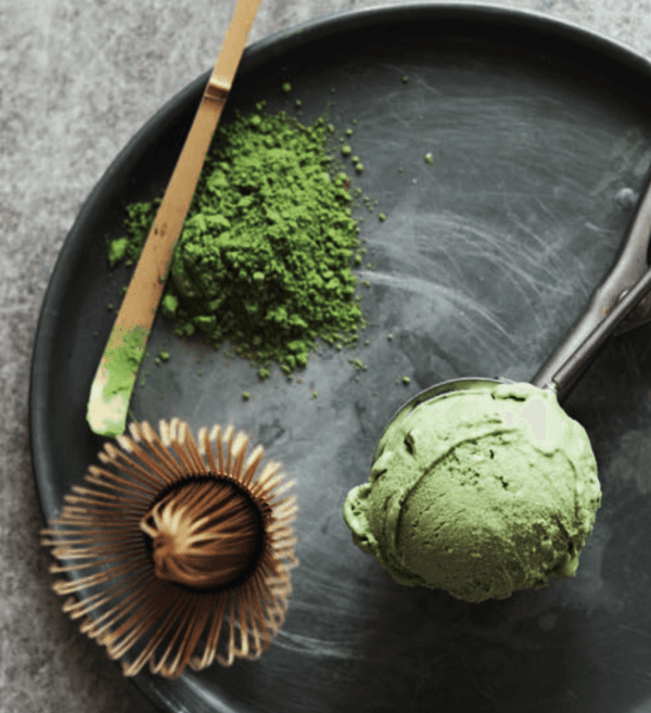 matcha-glace-gruentee-bio Auf einem schwarzen Teller liegen eine Kugel grünes Matcha-Eis, ein Bambusbesen, ein Löffel und Matcha-Pulver – ideal für alle, die Matcha Pulver Bio kunstvoll arrangiert kaufen möchten.