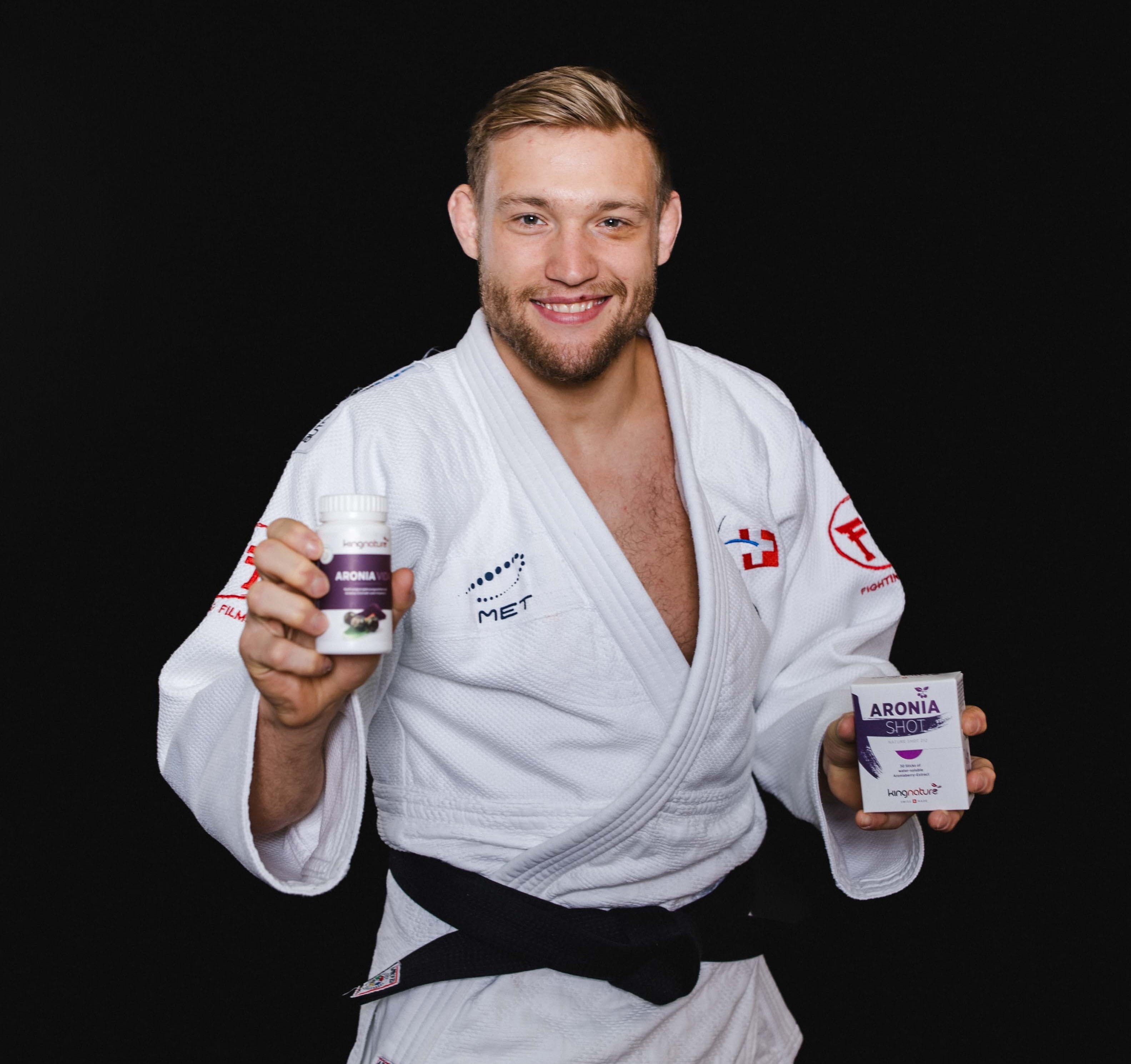 Ein Mann in einem weißen Judoanzug und mit schwarzem Gürtel lächelt, während er vor einem schwarzen Hintergrund eine Flasche und eine Schachtel Aronia-Nahrungsergänzungsmittel hält und die Kraft von Aronia demonstriert.