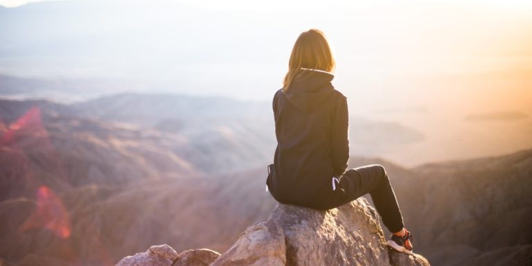 Eine Person in einer schwarzen Jacke sitzt auf einem Felsen und überblickt eine weite Alpenlandschaft bei Sonnenaufgang oder Sonnenuntergang, wobei sanftes Sonnenlicht die ruhige Szene erhellt.