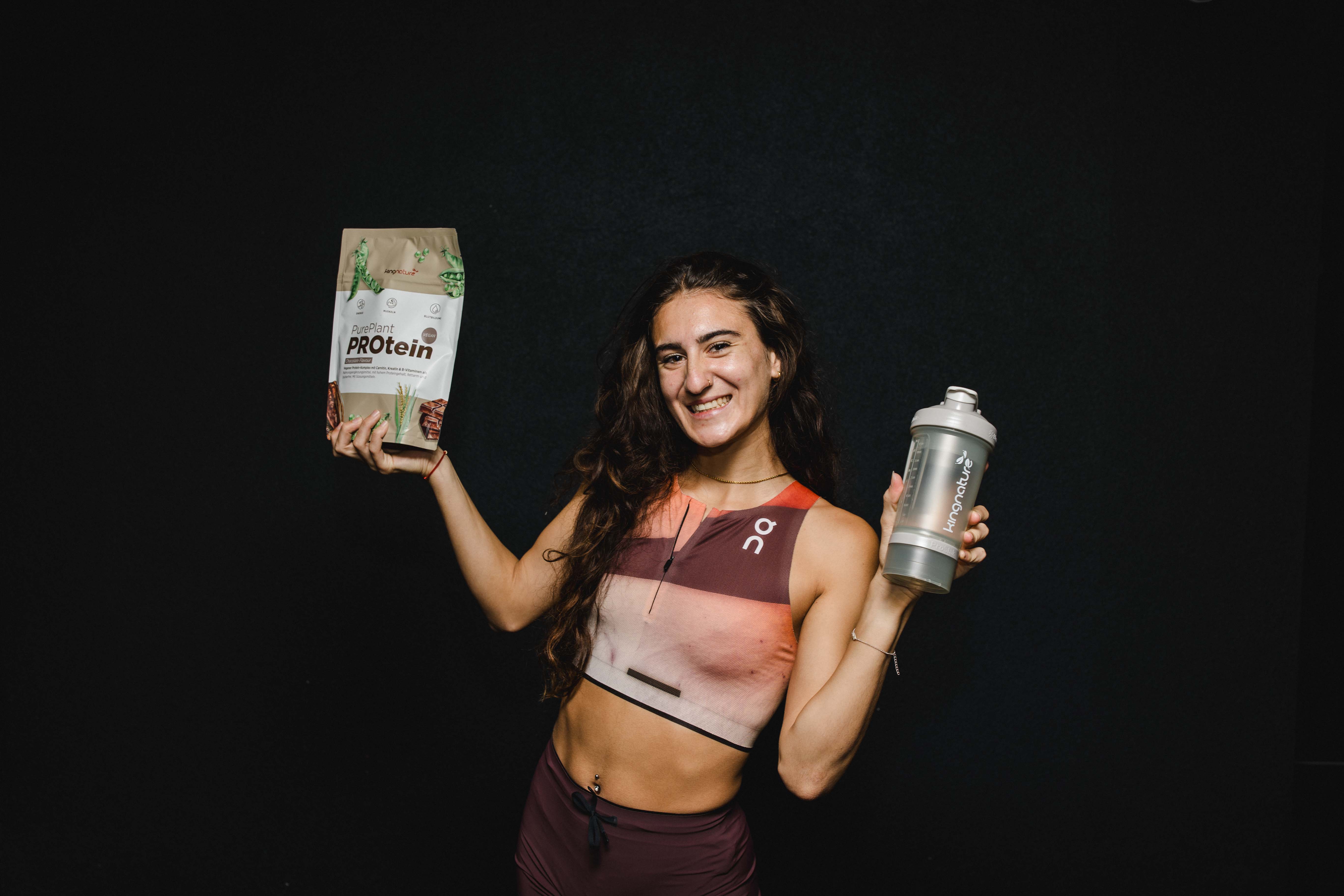 Valentina Rosamilia lächelt in Sportkleidung, hält in der einen Hand eine Tüte veganes Proteinpulver und in der anderen eine Shaker-Flasche und steht vor einem schlichten schwarzen Hintergrund.