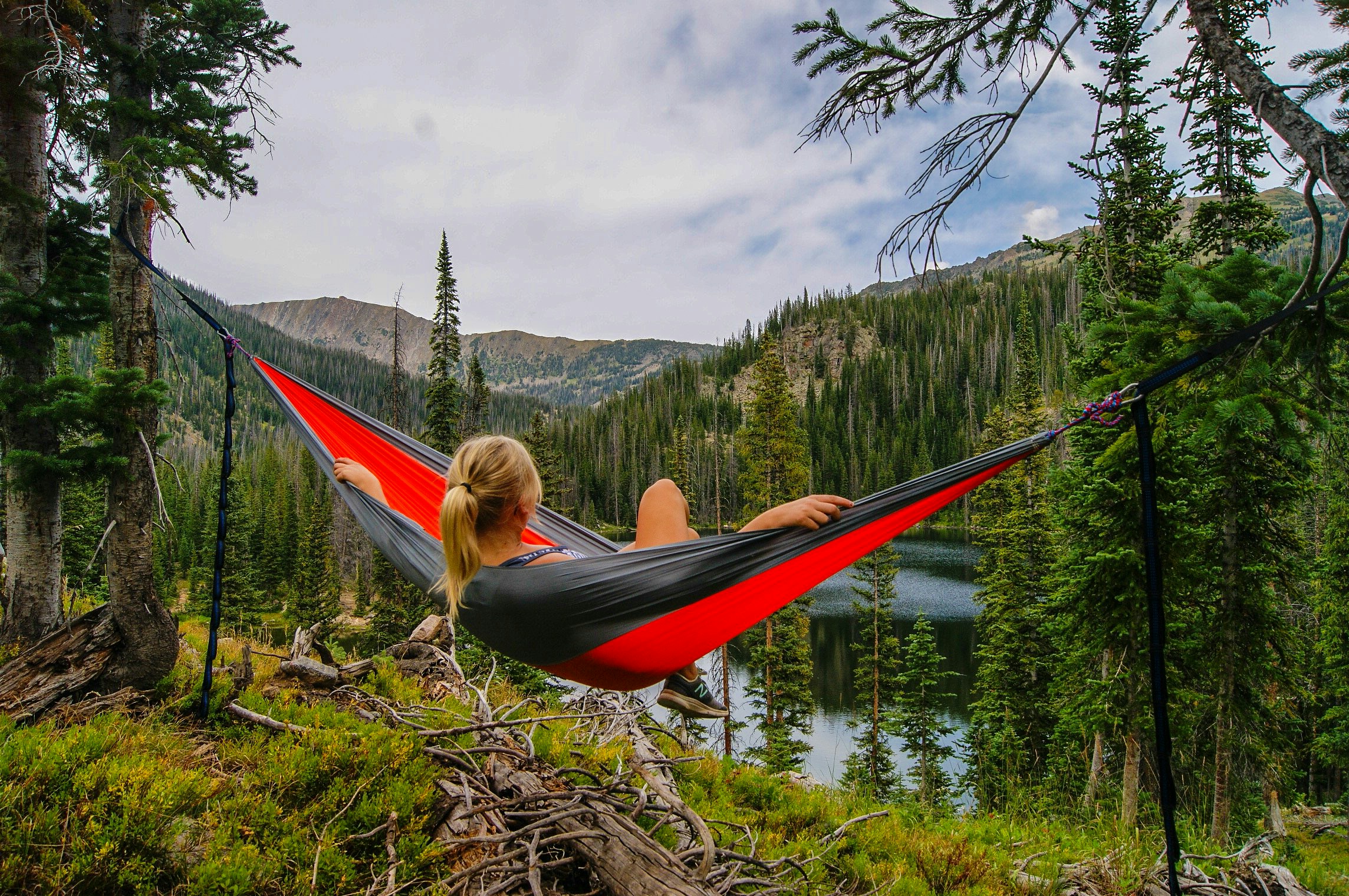 Eine Person mit blondem Haar entspannt sich in einer rot-grauen Hängematte, die zwischen Bäumen aufgespannt ist, und genießt die Sommerzeit mit Blick auf eine bewaldete Berglandschaft mit einem See unter einem teilweise bewölkten Himmel.